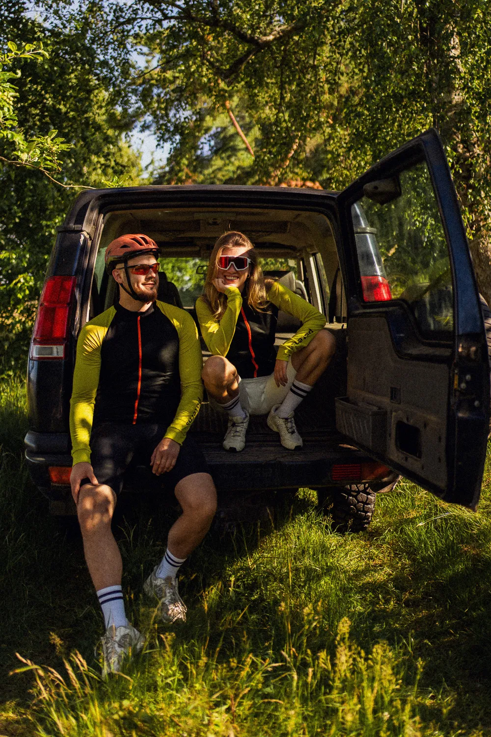 Dwóch rowerzystów odpoczywa w bagażniku SUV-a w skąpanym w słońcu lesie, uśmiechając się. Obaj mają na sobie identyczne kaski, okulary przeciwsłoneczne oraz czarną męską koszulkę rowerową z wełny merynosowej z długim rękawem: Koszulka rowerowa z wełny merynosowej z długim rękawem, męska, czarna.