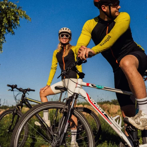 Dwóch rowerzystów w kaskach, okularach przeciwsłonecznych i identycznych czarnych męskich koszulkach rowerowych z wełny merino z długim rękawem zatrzymuje się na rowerach na trawie pod gołym niebem, pod czystym, błękitnym niebem.
