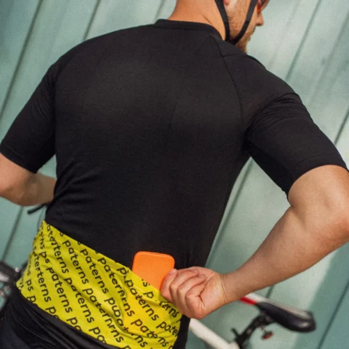 Rowerzysta ubrany w koszulkę rowerową z wełny merynosowej z krótkim rękawem dla mężczyzn w kolorze czarnym oraz w różowym kasku stoi obok białego roweru, sięgając do żółtej kieszeni koszulki i trzymając w prawej ręce pomarańczowy przedmiot.