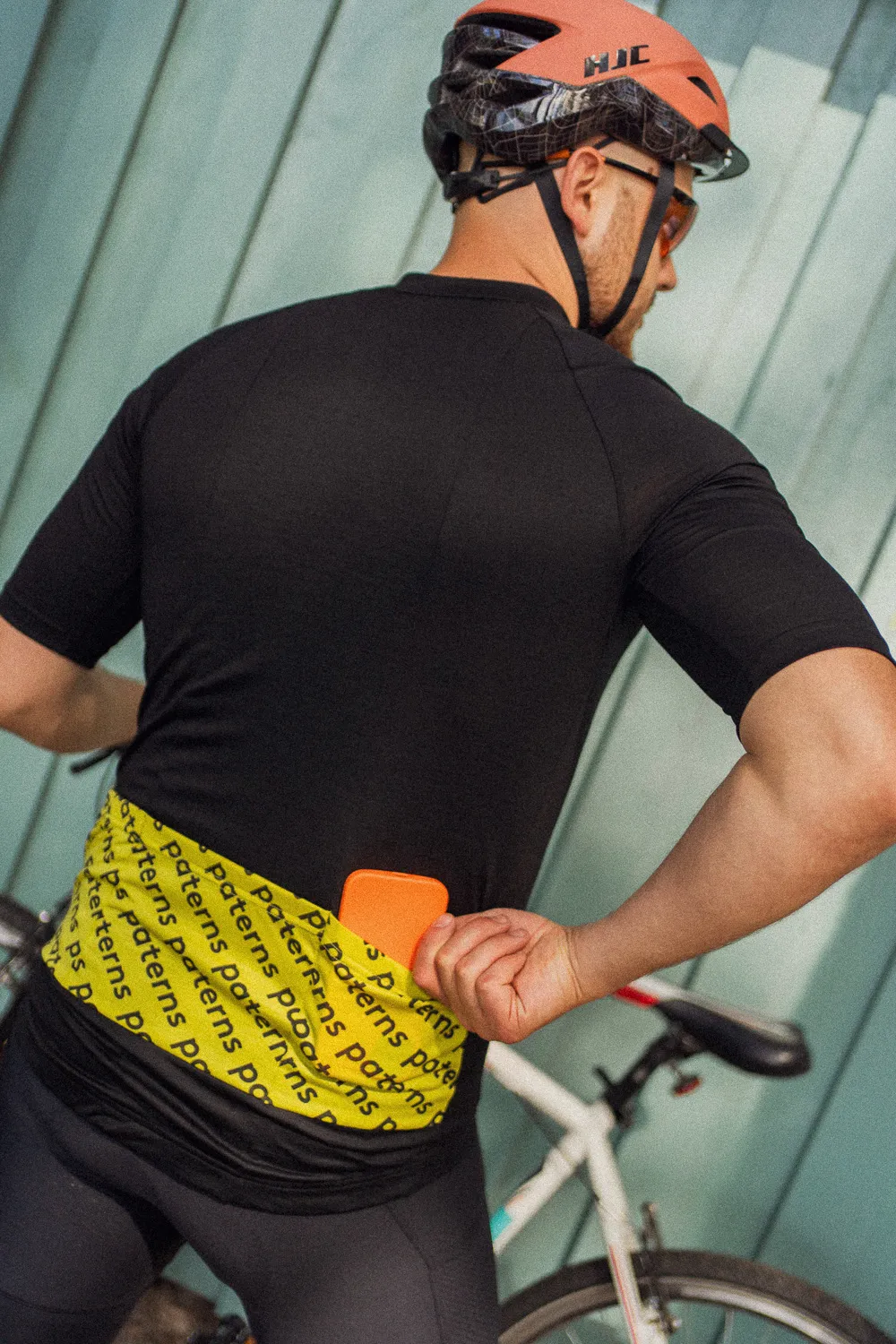 Rowerzysta ubrany w koszulkę rowerową z wełny merynosowej z krótkim rękawem dla mężczyzn w kolorze czarnym oraz w różowym kasku stoi obok białego roweru, sięgając do żółtej kieszeni koszulki i trzymając w prawej ręce pomarańczowy przedmiot.