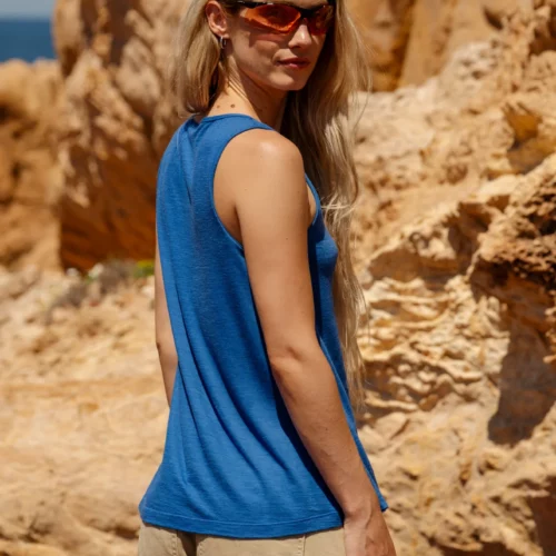 Kobieta z długimi blond włosami i okularami przeciwsłonecznymi, ubrana w Top z wełny merino flare damski kobalt i beżowe spodnie, stoi na zewnątrz przed dużymi, oświetlonymi słońcem formacjami skalnymi.