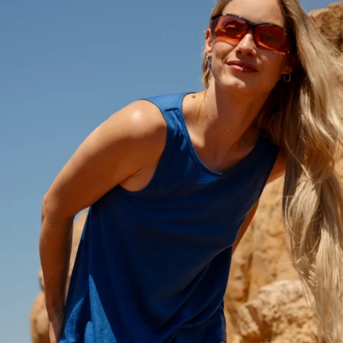 Kobieta z długimi blond włosami, czerwonymi okularami przeciwsłonecznymi i beżowymi szortami nosi Top z wełny merino flare damski kobalt, pochylając się do przodu i uśmiechając się na zewnątrz na tle opalonych formacji skalnych pod czystym błękitnym niebem.