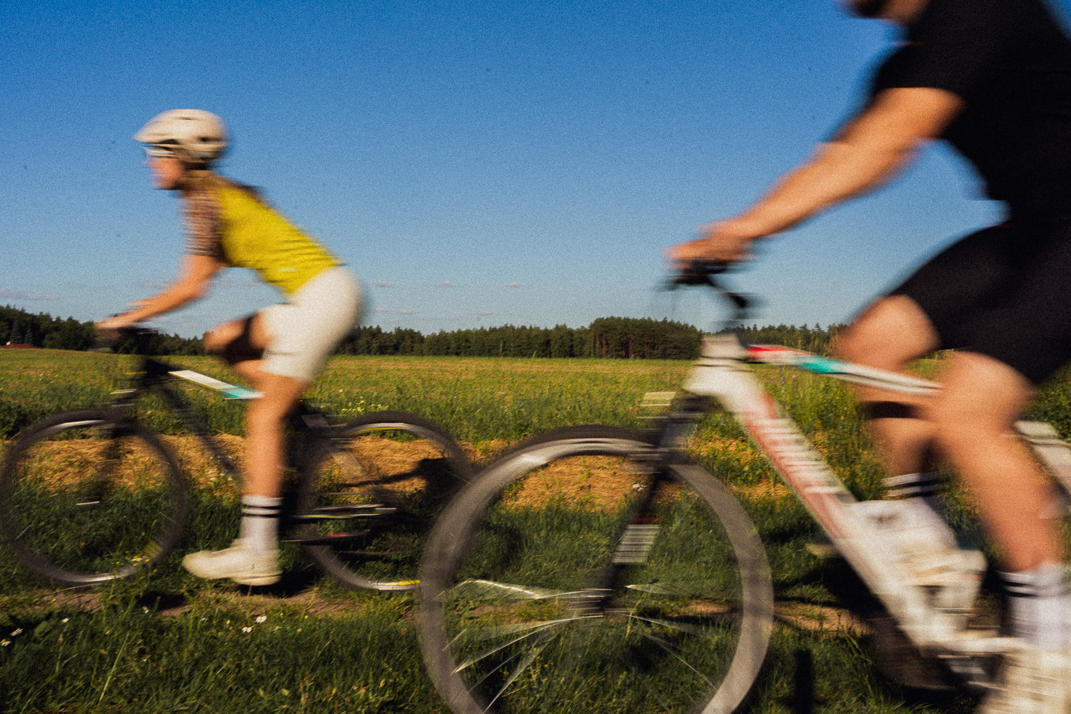 Dwóch rowerzystów pędzi po wiejskiej ścieżce, a ich sylwetki są rozmyte w ruchu; otaczają ich zielone pola i czyste, błękitne niebo. Obaj mają na sobie kaski i stroje sportowe. W oddali widać drzewa.