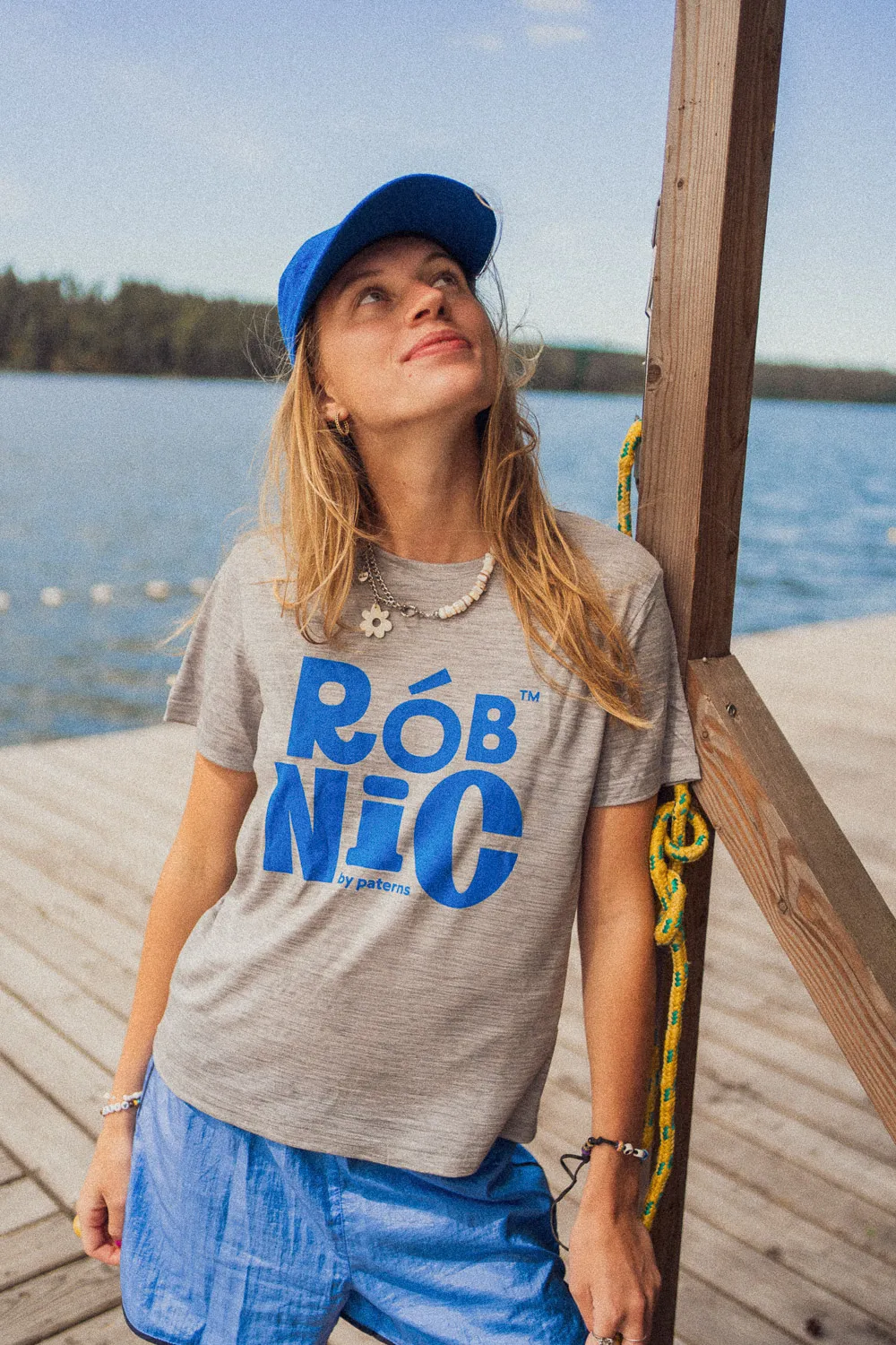 Kobieta w niebieskiej czapce i niebieskich szortach opiera się o drewniany słupek przy pomostu nad jeziorem. Ma na sobie szarą koszulkę z wyrazistym niebieskim napisem „RÓB NIC” i spogląda w górę, lekko się uśmiechając. W tle widać las i wodę.