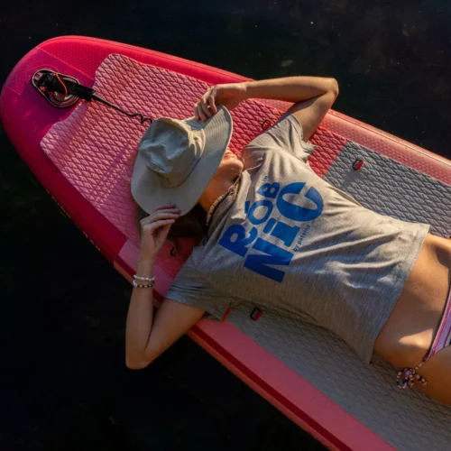 Osoba ubrana w szarą koszulkę i bikini w paski odpoczywa na różowej desce do paddleboardingu, trzymając kapelusz przeciwsłoneczny nad twarzą, leżąc na plecach na tle ciemnej wody.