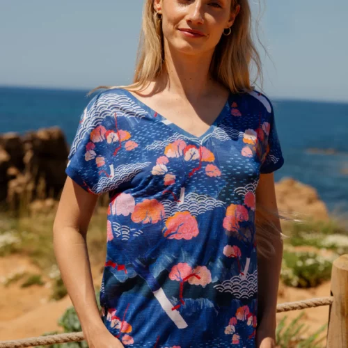 Kobieta o długich blond włosach stoi na świeżym powietrzu nad brzegiem oceanu. Ma okulary przeciwsłoneczne założone na głowę, niebieską koszulkę w kolorowe kwiatowe wzory i fale oraz beżowe szorty. Niebo jest bezchmurne i słoneczne.