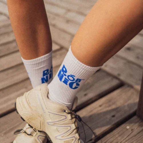 Zbliżenie na osobę stojącą na drewnianym tarasie, ubraną w beżowe trampki i białe skarpetki z niebieskim napisem „BE NICE”.