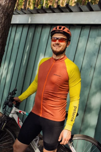 Mężczyzna ubrany w koszulkę rowerową z wełny merynosowej z długim rękawem w kolorze ceglastym, w kasku i okularach przeciwsłonecznych, stoi obok białego roweru przed niebiesko-zielonym drewnianym płotem i uśmiecha się do aparatu.