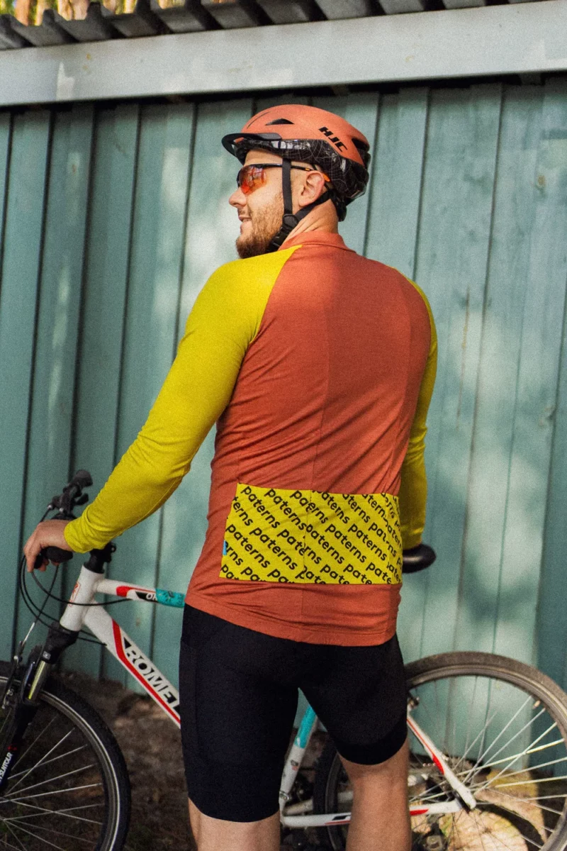 Rowerzysta w kasku, okularach przeciwsłonecznych i męskiej koszulce rowerowej z wełny merynosowej w kolorze ceglastym, z długimi rękawami, stoi obok białego roweru, odwrócony plecami do turkusowego drewnianego ogrodzenia.