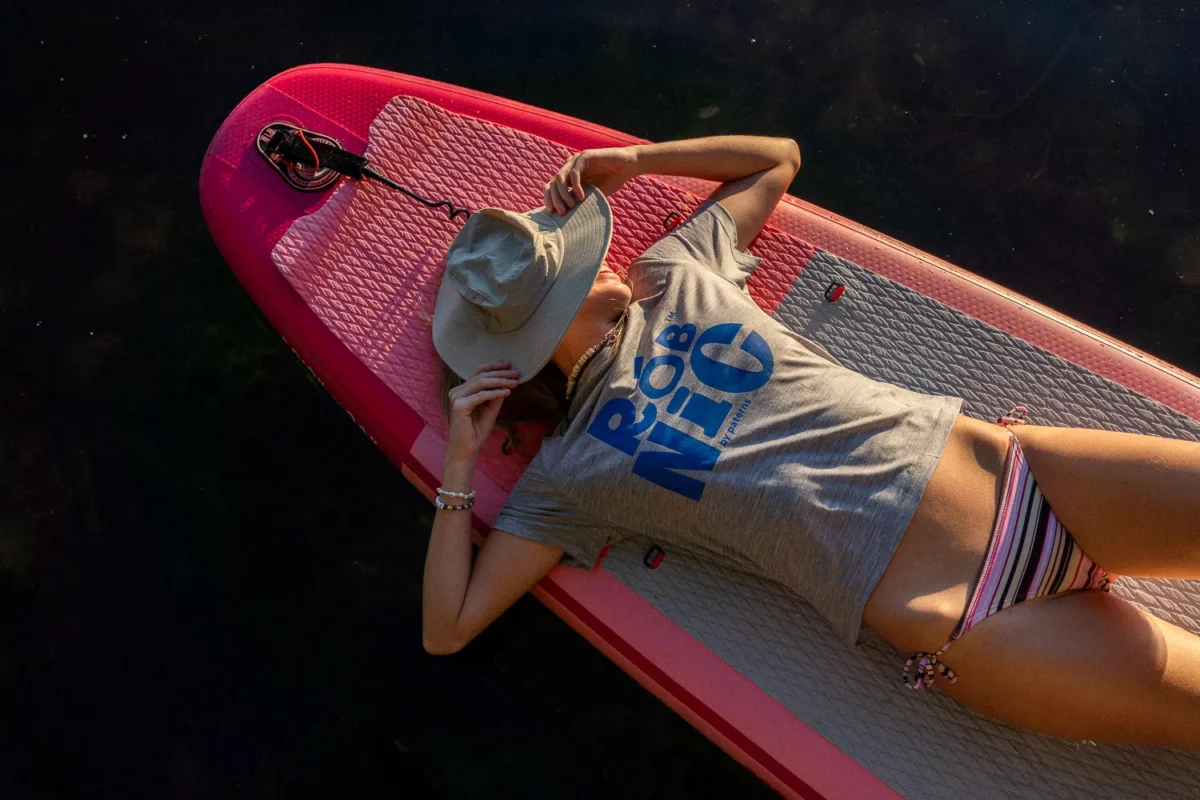 Osoba ubrana w szarą koszulkę i bikini w paski odpoczywa na różowej desce do paddleboardingu, trzymając kapelusz przeciwsłoneczny nad twarzą, leżąc na plecach na tle ciemnej wody.