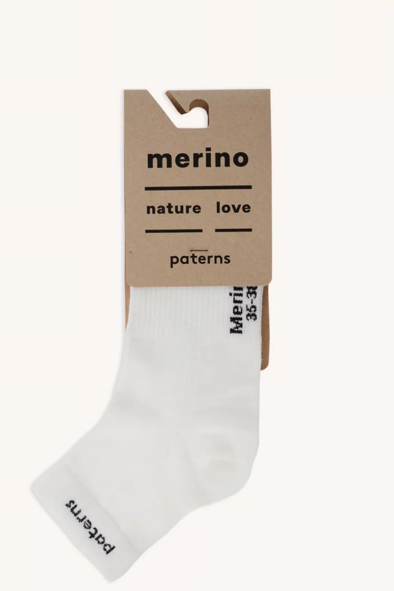 Pojedyncza biała skarpeta przymocowana do brązowej kartonowej etykiety z napisem „merino”, „nature love” i „paterns”. Na skarpecie widnieje czarny napis w okolicy palców oraz wzdłuż ściągacza, dzięki czemu idealnie pasuje do czapki z wełny merino z ściągaczem w kolorze różowym melanżowym.
